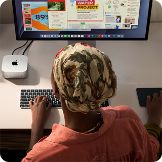 Човек пише на бюро с Mac mini, свързан със Studio Display, Magic Keyboard, Magic Trackpad, различни неща