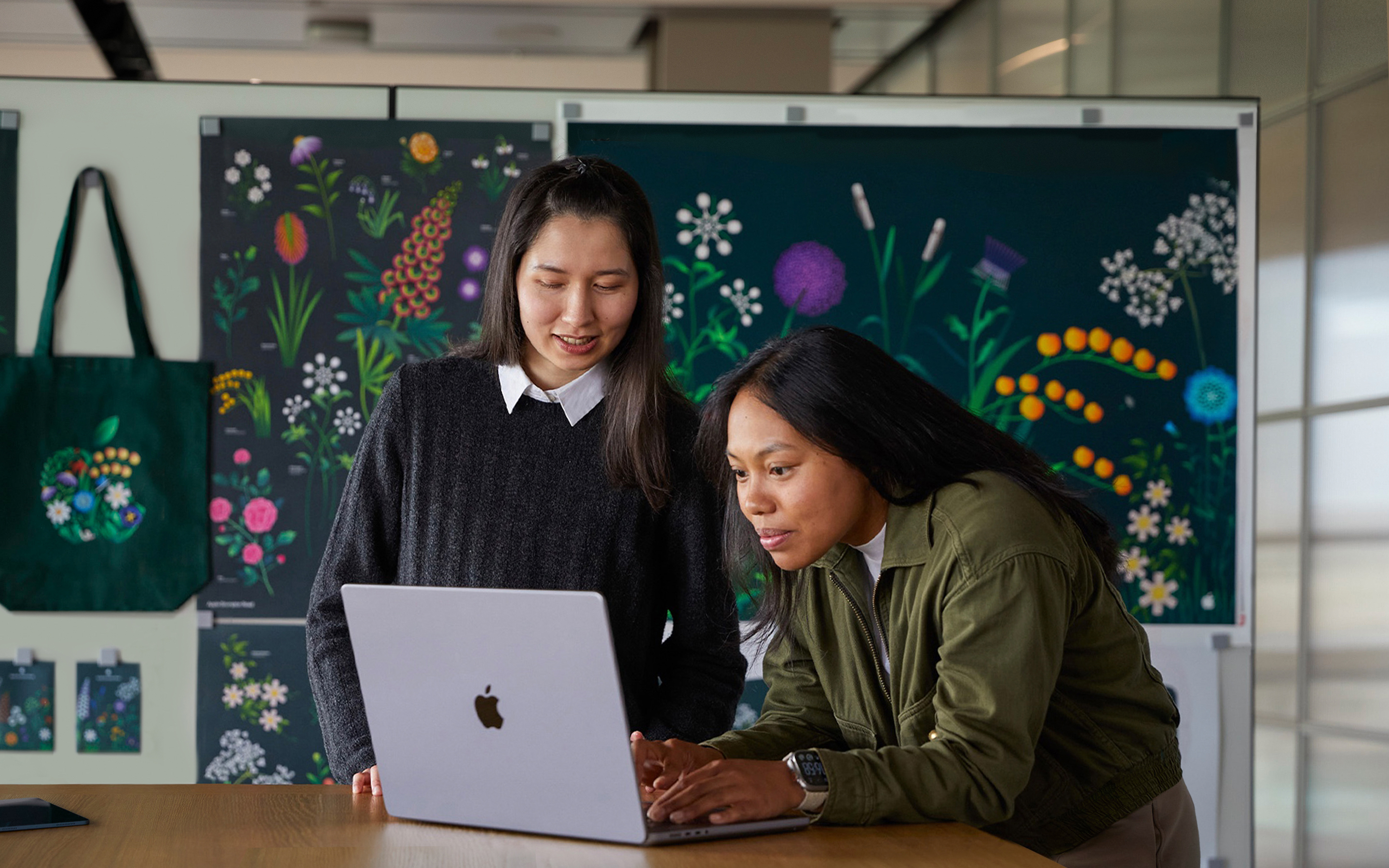 兩位 Apple 市場推廣團隊成員在其團隊設計的海報前，一起使用 MacBook 工作。