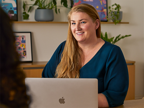 Een Apple medewerker die in een algemene ruimte aan een tafel achter een MacBook zit en met een collega praat.
