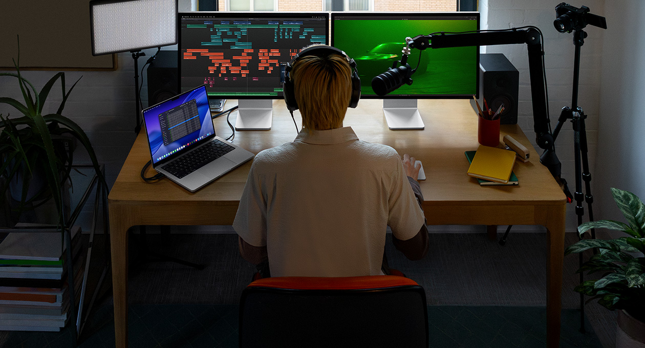 Seseorang sedang duduk di depan meja dengan mikrofon dan headphone di depan tiga perangkat, layar Mac Studio Display, alat pengeditan audio, klip suara dalam berbagai blok warna berbeda, layar Mac Studio Display kedua menampilkan foto mobil dalam kabut dengan pencahayaan hijau bernuansa sentimental, layar MacBook Pro 16 menampilkan aplikasi Finder di desktop yang memperlihatkan File untuk Final Cut Pro, dock, latar belakang desktop bergaya biru