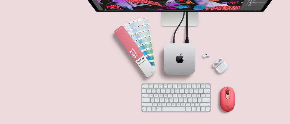 Skats no augšas uz Mac mini sudraba krāsas alumīnija korpusā ar noapaļotiem stūriem un spīdīgu melnu Apple logotipu centrā, savienots ar vadu. Augšpusē daļēji redzams Studio Display, atvērti AirPod 4, Pantone krāsu paletes, cipartastatūra un pele