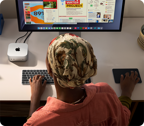 O persoană tastând la un birou cu un Mac mini, Studio Display conectat, Magic Keyboard, Magic Trackpad, diverse obiecte