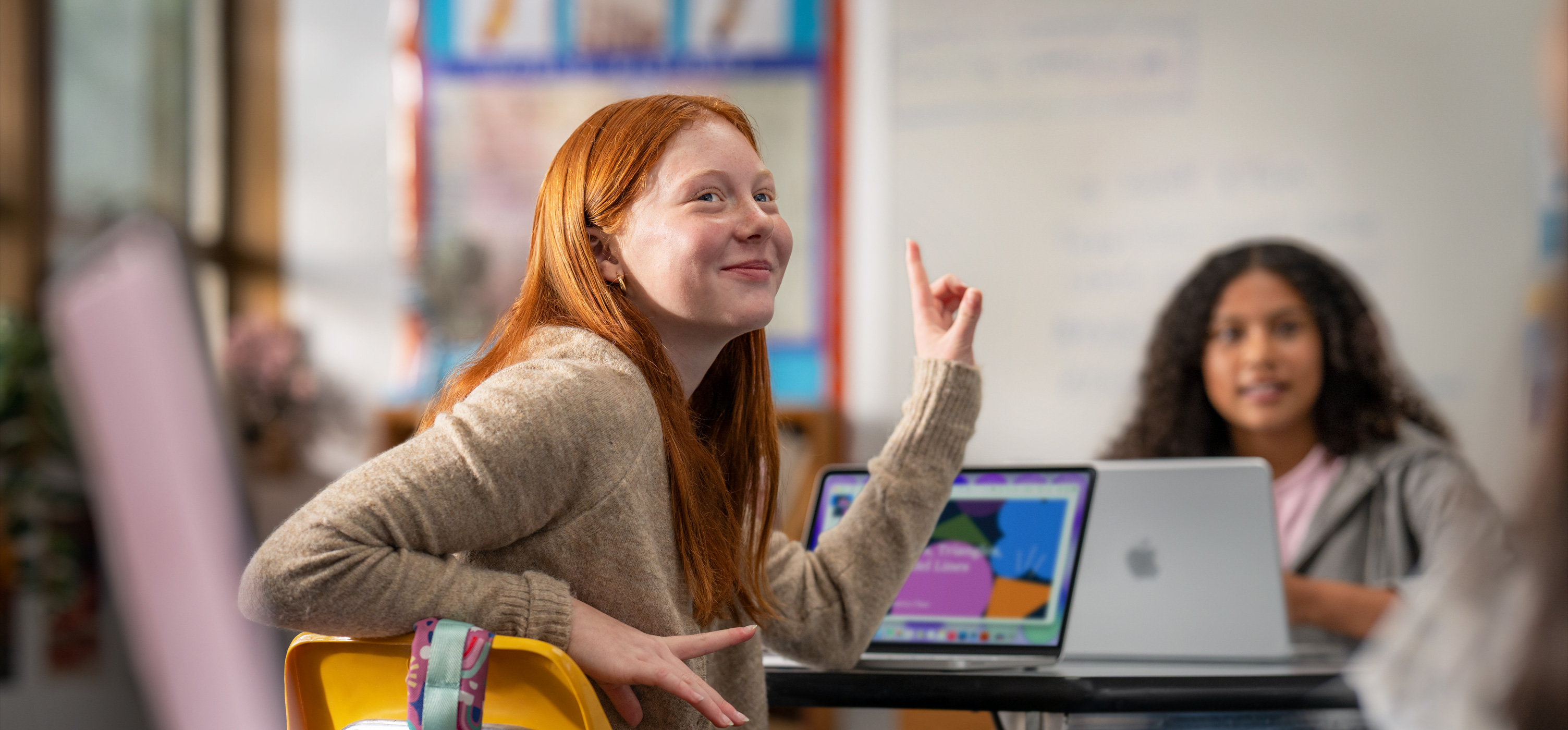 Estudante sentada em frente a um MacBook na sala de aula levanta a mão para responder a uma pergunta, sugerindo facilidade de uso e envolvimento