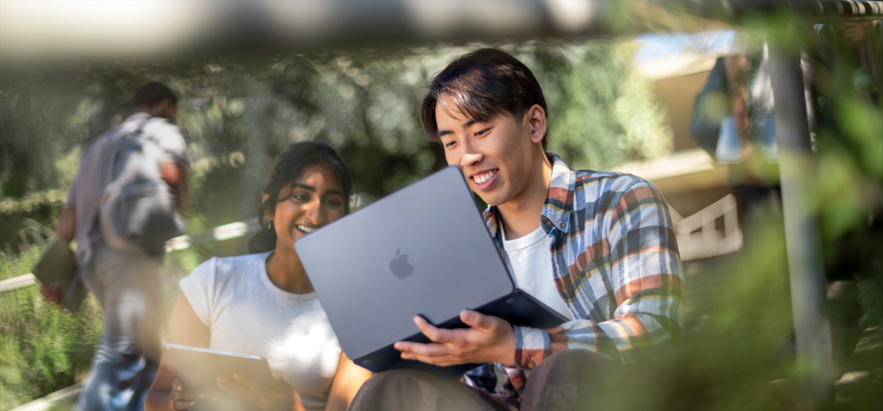 Dois estudantes sorridentes usam um MacBook ao ar livre. Um deles segura o notebook no colo enquanto colaboram em uma tarefa