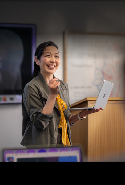 Uma professora faz uma apresentação para sua turma em um auditório enquanto segura um MacBook prateado, demonstrando o desempenho e a bateria para o dia todo