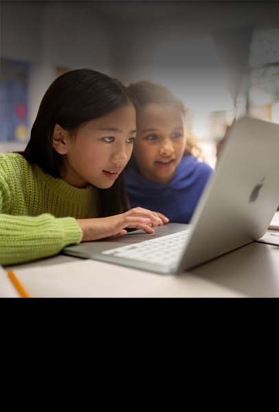 Duas jovens estudantes sentadas a uma mesa observam atentamente um MacBook enquanto colaboram em sala de aula