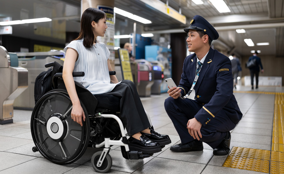 Une femme en fauteuil roulant, habillée de manière professionnelle, discute avec un agent de station sur le quai.