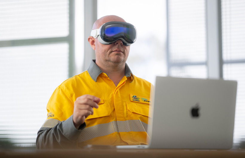 En medarbetare som bär Apple Vision Pro gör en gest med fingrarna i luften, medan en MacBook Pro i silver står öppen på skrivbordet
