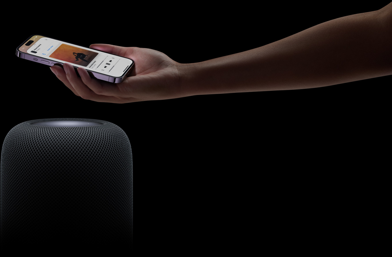 Een hand die van rechts het beeld in komt en een iPhone boven een HomePod-speaker houdt