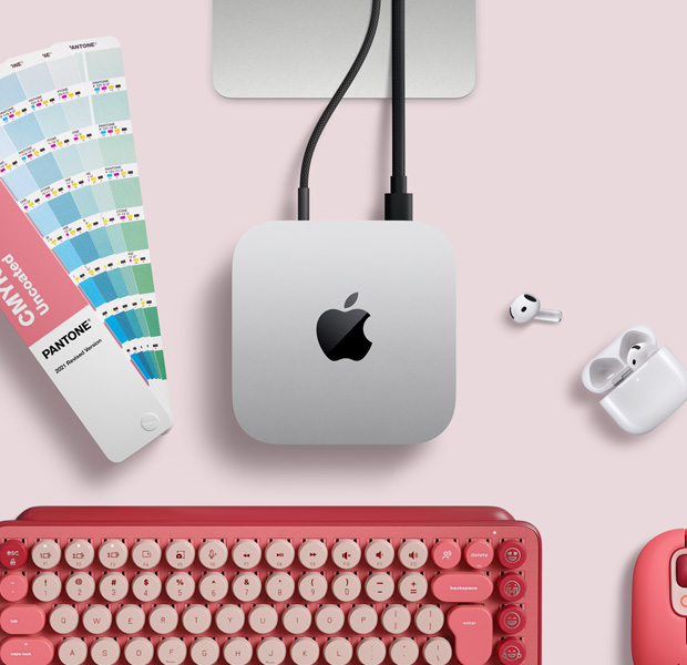 Mac mini, dessus, aluminium argent, angles arrondis, logo Apple noir brillant centré, connecté par un câble, support du Studio Display partiellement visible, AirPods 4, pastilles Pantone, clavier et souris Logitech rouges