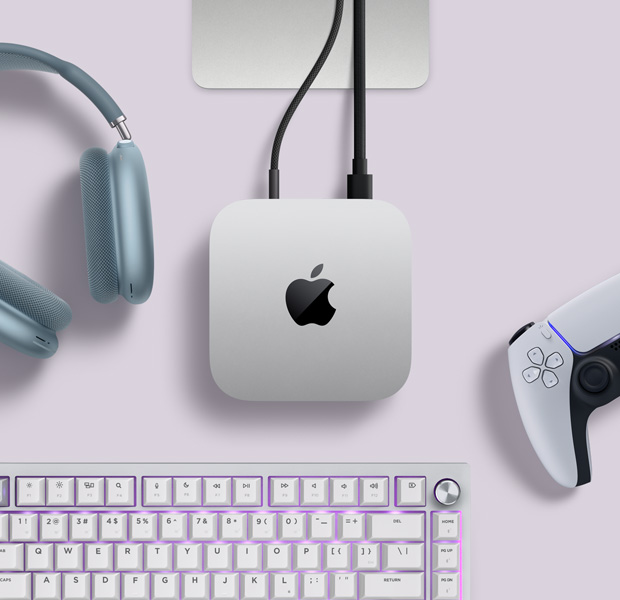 Mac mini, dessus, aluminium argent, angles arrondis, logo Apple noir brillant centré, connecté par un câble, support du Studio Display partiellement visible, AirPods Max 2, couleur bleue, manette PlayStation DualSense, couleur blanche, et clavier blanc